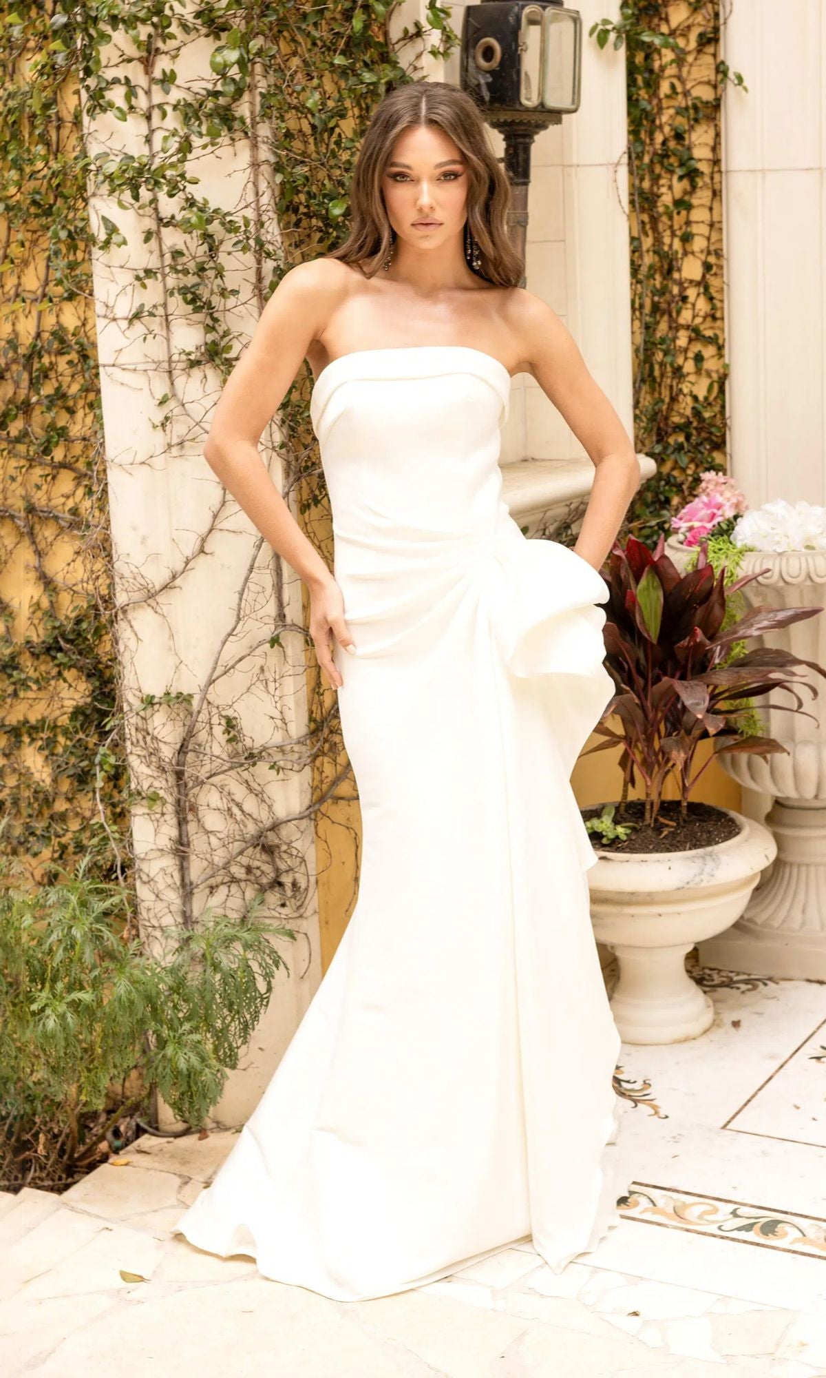 A woman in an ivory Terani 2111P4019 long formal dress with a strapless neckline and bow detail stands outdoors near potted plants and an ivy-covered wall.